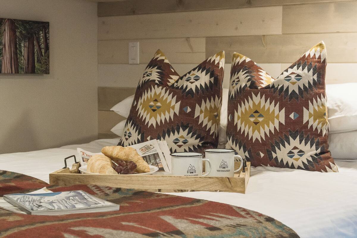 A bed with patterned pillows, a tray with croissants and a mug, and a wall with a painting.