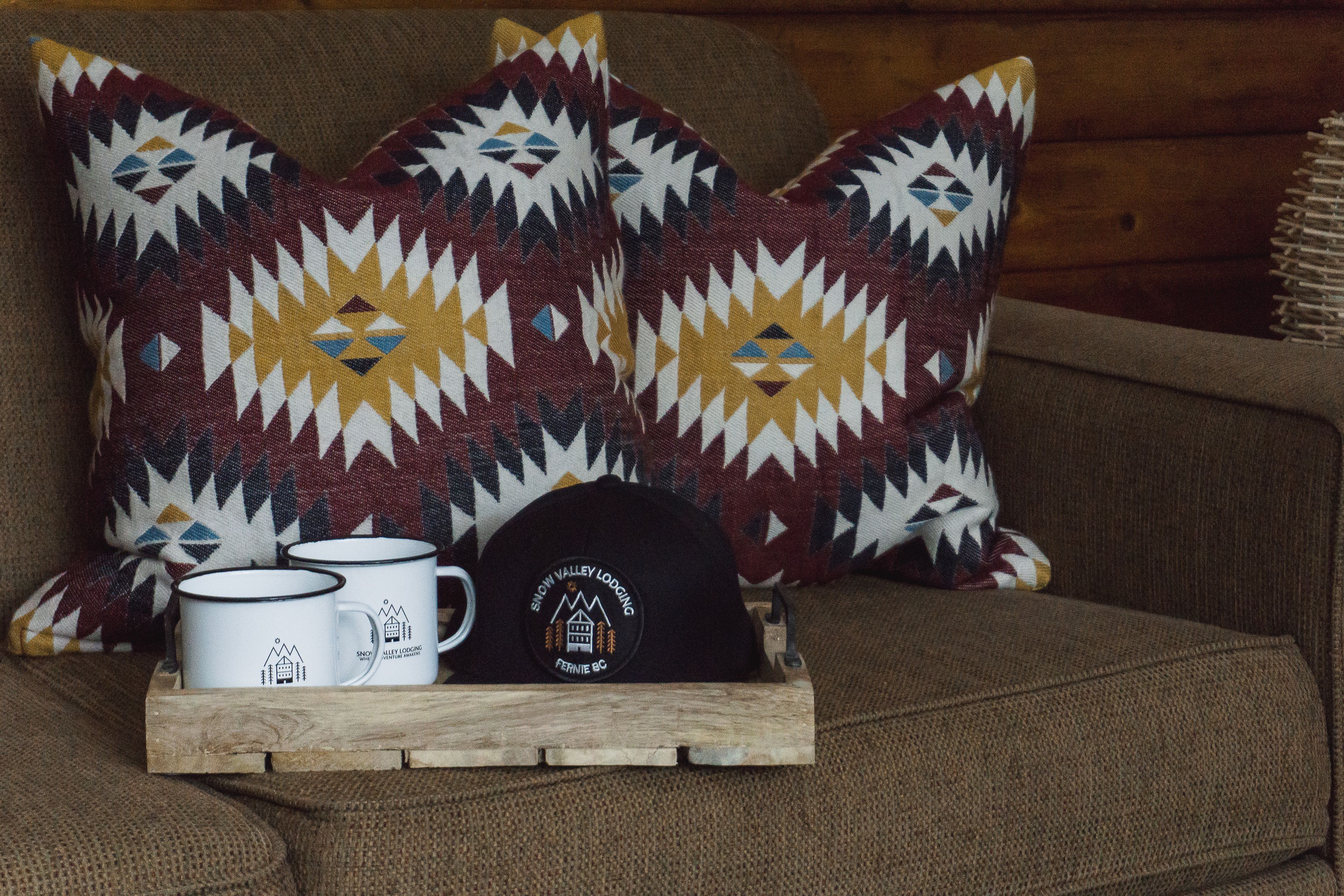 Un plateau en bois avec deux tasses et une casquette noire, posé sur un canapé agrémenté de coussins à motifs.