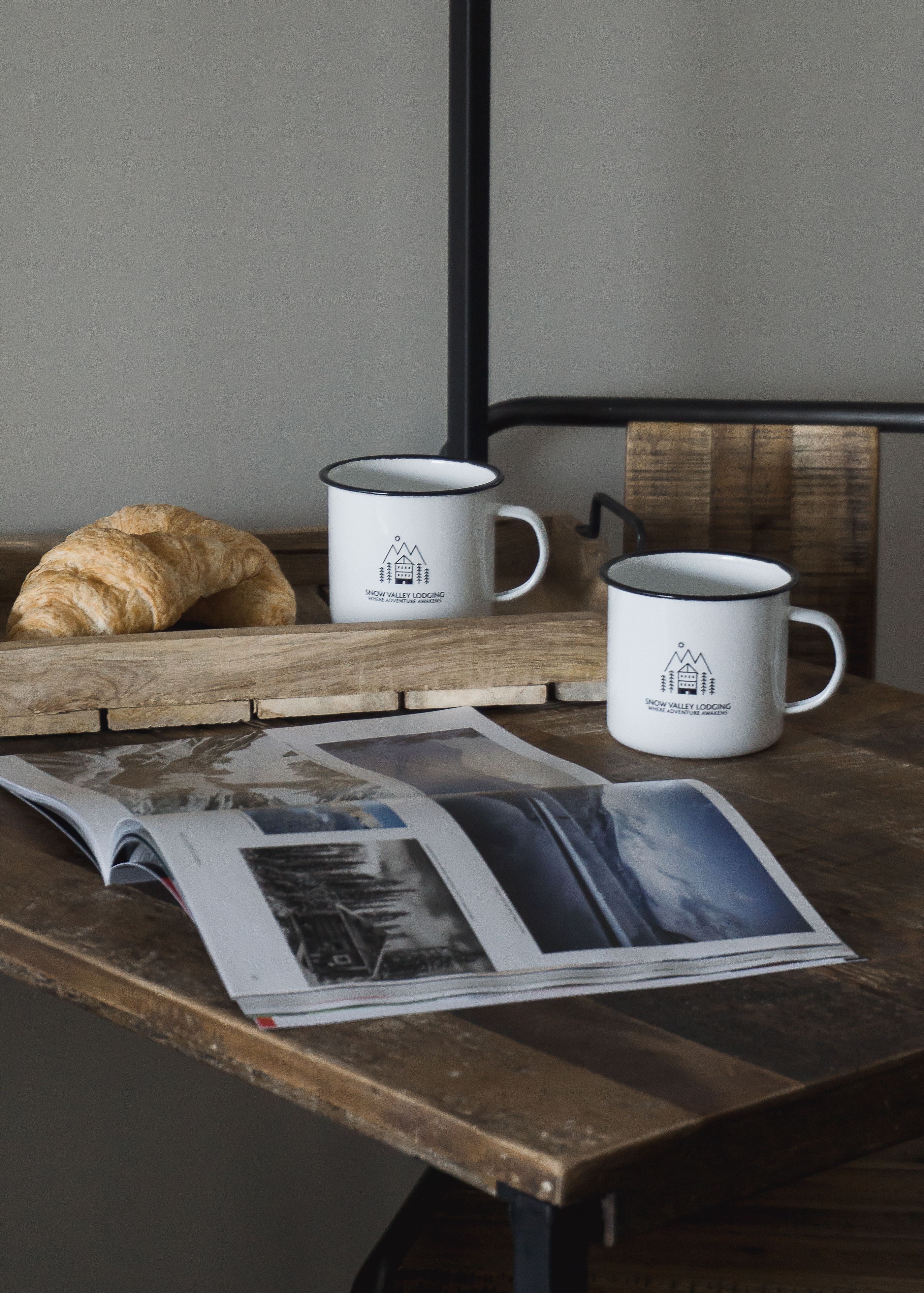Une table en bois sur laquelle sont posées deux tasses blanches ornées d’un logo représentant une montagne et un chalet, un croissant et un magazine contenant des photos de voyage.