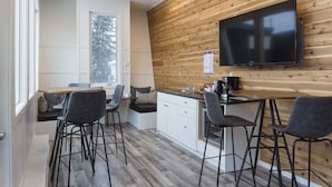 A modern kitchen with wooden paneling, a large flat-screen TV, bar stools, and a window with a view of snow-covered trees. - Snow Valley Lodging (Fernie)