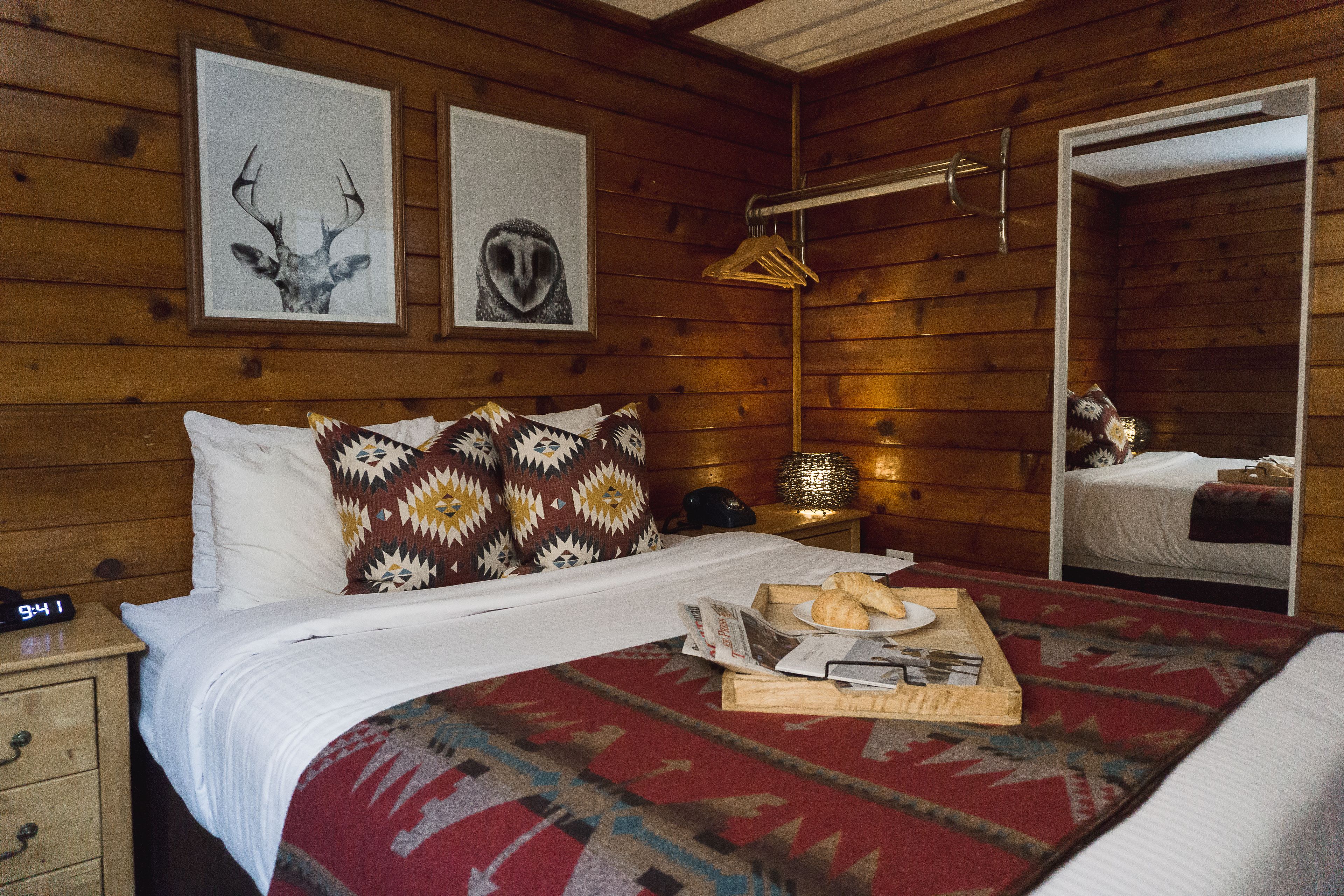 Un dormitorio de cabaña de madera con cama, una mesita de noche, un espejo y dos cuadros en la pared.