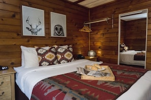 A wooden cabin bedroom with a bed, a bedside table, a mirror, and two framed pictures on the wall.