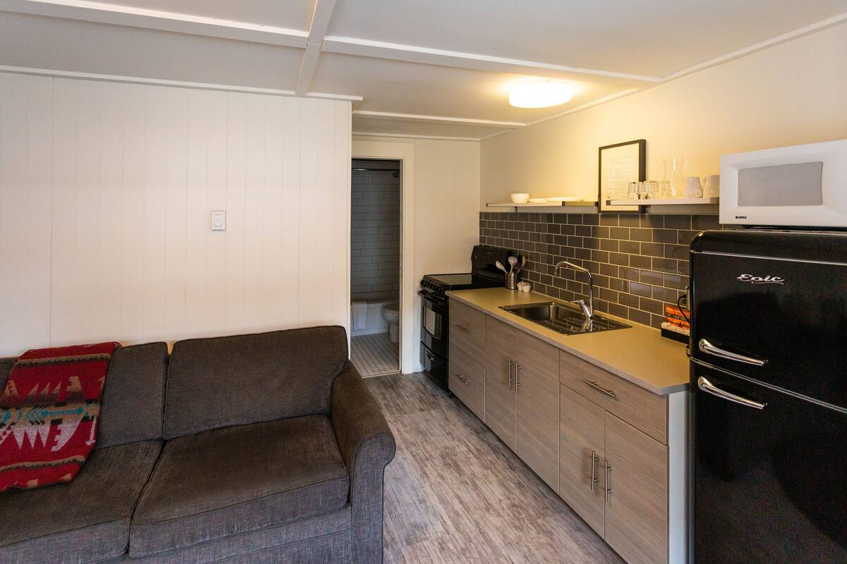 A compact kitchen with a refrigerator, stove, sink, and microwave. There is a sofa with a blanket and a door leading to a bathroom.