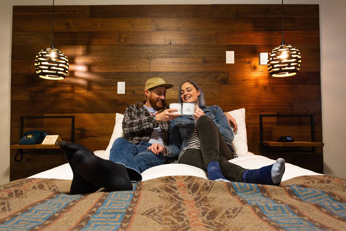 A couple on a bed with patterned bedding, wooden headboard, and pendant lights.