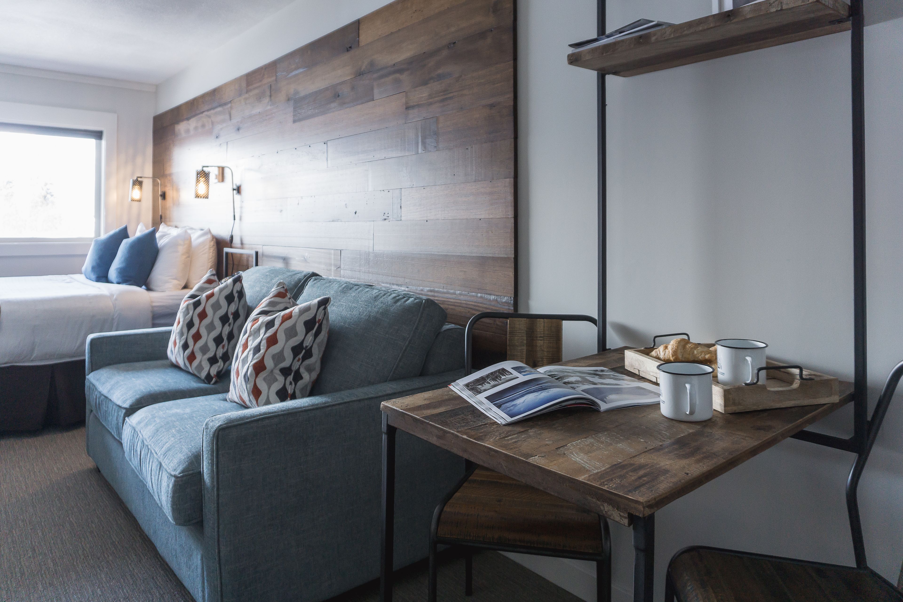 A cozy room with a wooden headboard, a grey sofa with patterned cushions, a wooden table with a magazine, two mugs, and a tray with croissants.
