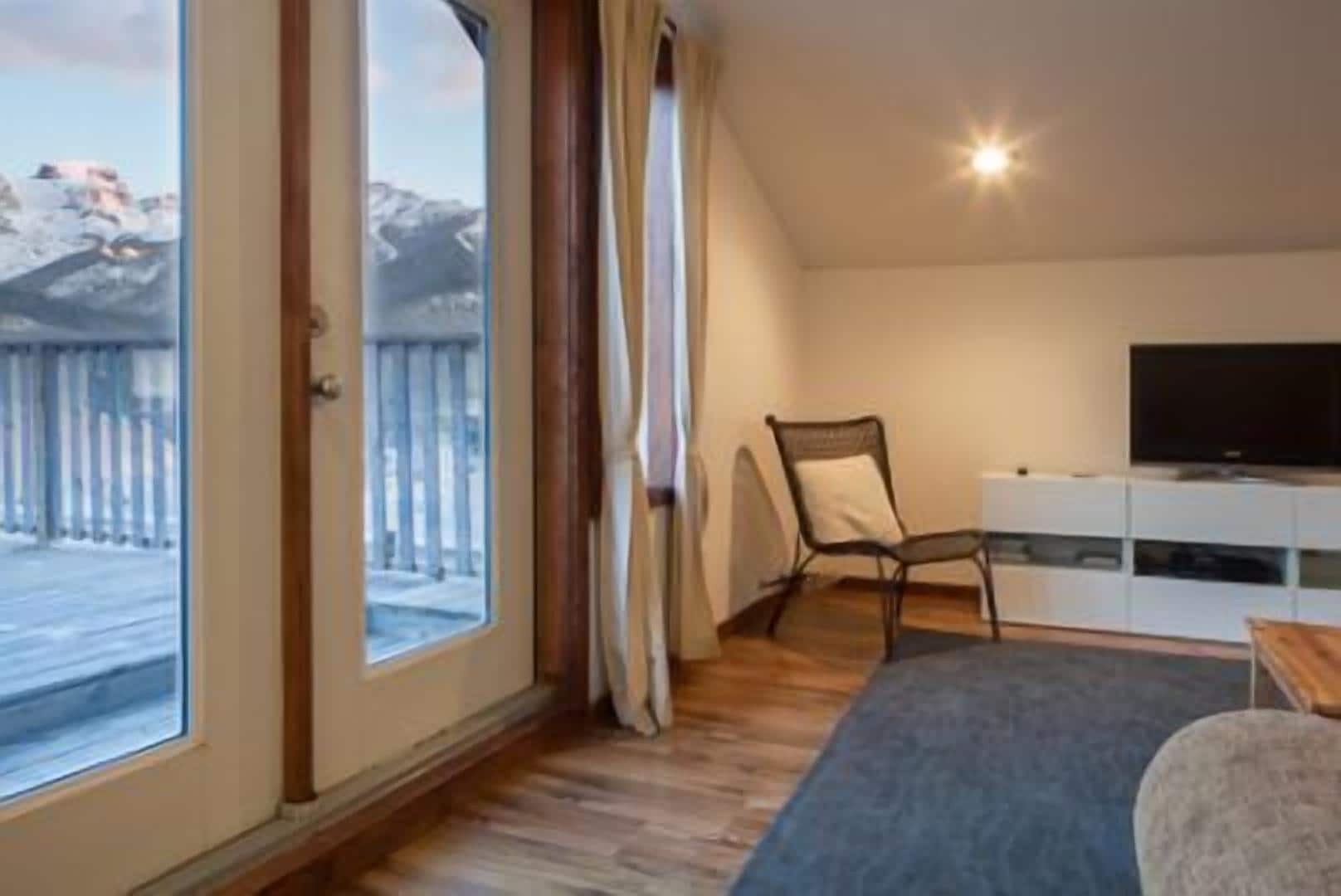 A room with a large sliding glass door leading to a snowy outdoor area, a television on a white cabinet, and a chair.