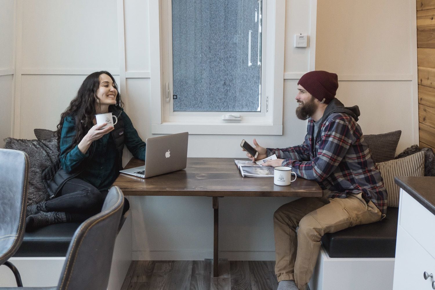 Two people sitting at a wooden table with a laptop and a mug, in a room with a window and a thermostat on the wall.