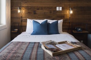 Una cama bien hecha con almohadas azules y blancas, un cabecero de madera y una bandeja con material de lectura sobre una colcha estampada.