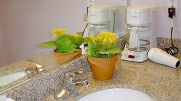 Um banheiro com bancada de granito, pia, duas cafeteiras, secador de cabelo e uma planta em vaso com flores amarelas.