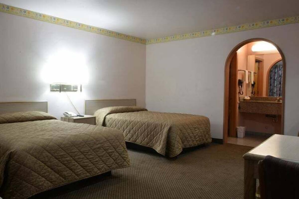 A hotel room with two beds, a desk, a chair, a nightstand, a lamp, and a bathroom visible through an archway.