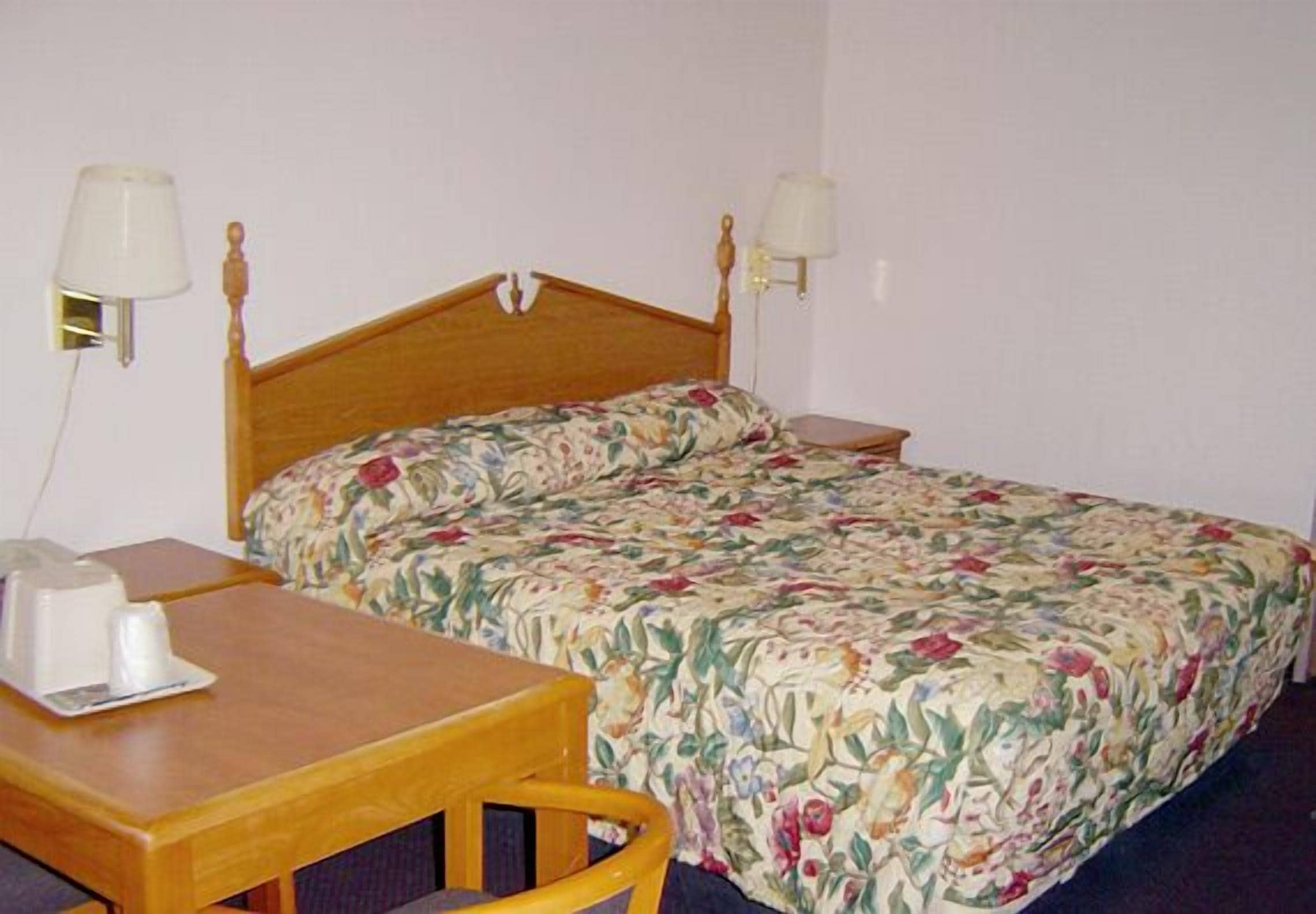 A single bed with a floral bedspread, a wooden nightstand, a bedside table with a lamp, and a wall-mounted lamp.