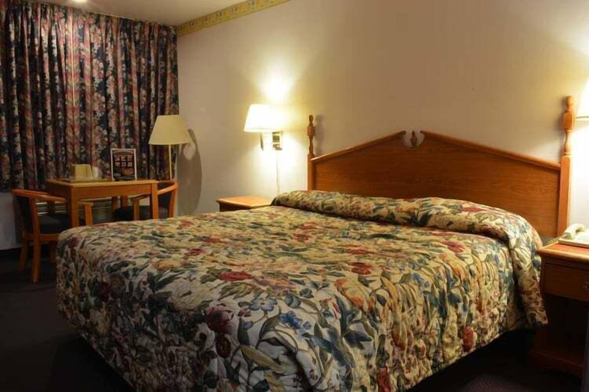 A hotel room with a floral-patterned bedspread, a wooden headboard, a bedside table with a lamp, and a desk with a chair.