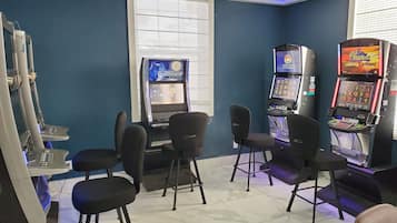 Zona de juegos con máquinas tragamonedas, una mesa con sillas y una pantalla grande mostrando un juego.