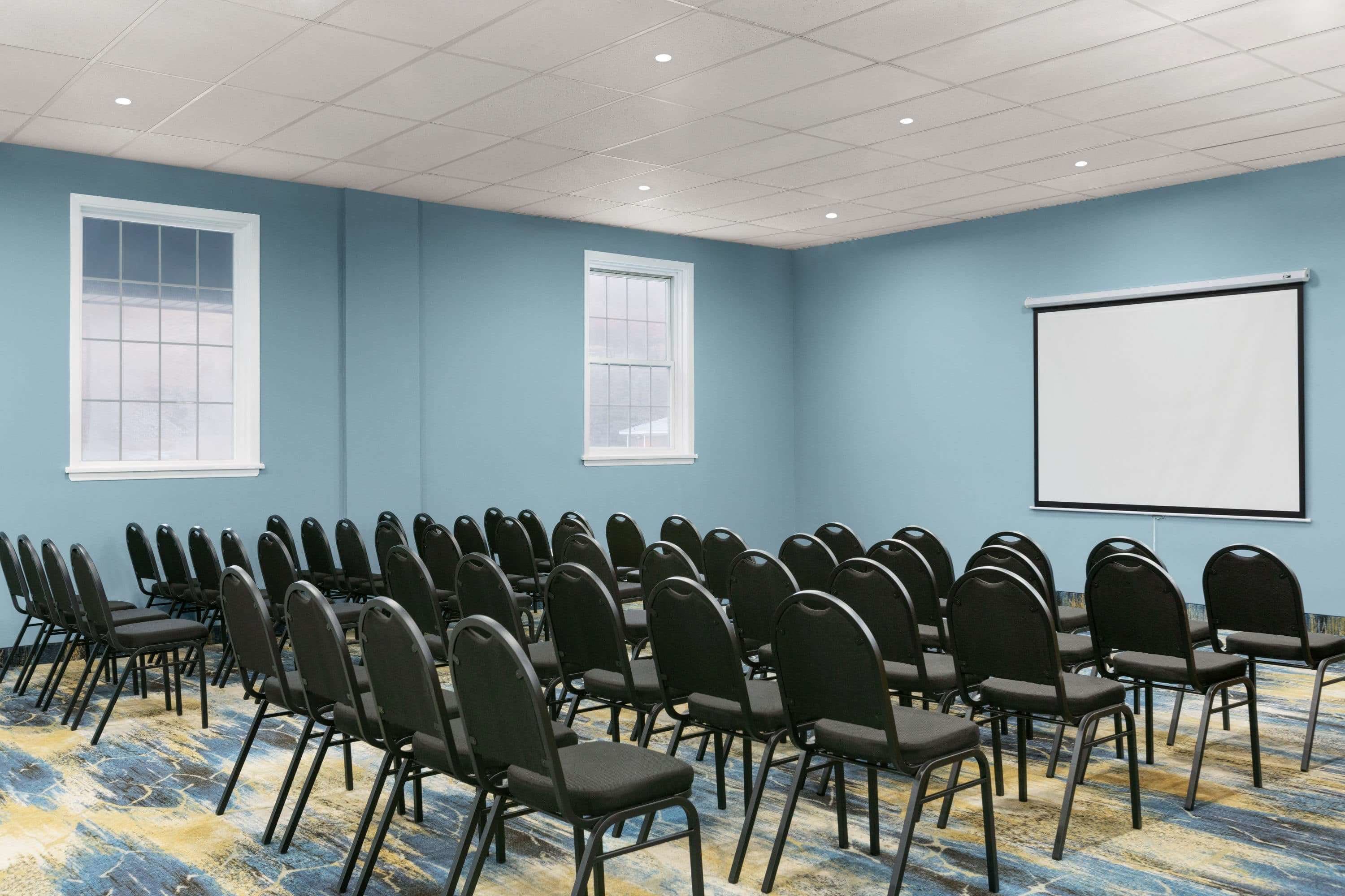 Uma sala de conferência com fileiras de cadeiras, uma tela de projetor e duas janelas.
