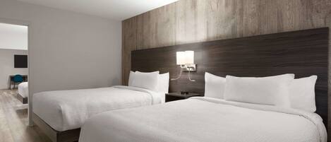 A hotel room with two beds, a wooden headboard, a bedside table with a lamp, and a view of the hallway.