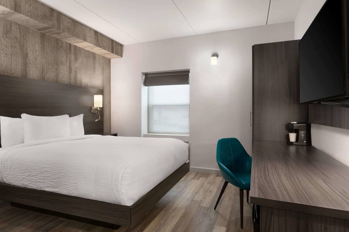 Ett modernt hotellrum med en stor säng, ett skrivbord med en stol, en TV och ett fönster med persienner.