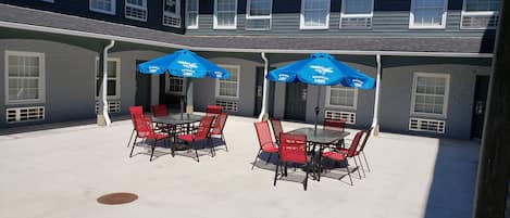 An outdoor patio with two tables and chairs, covered by blue umbrellas, and a building with multiple windows in the background.