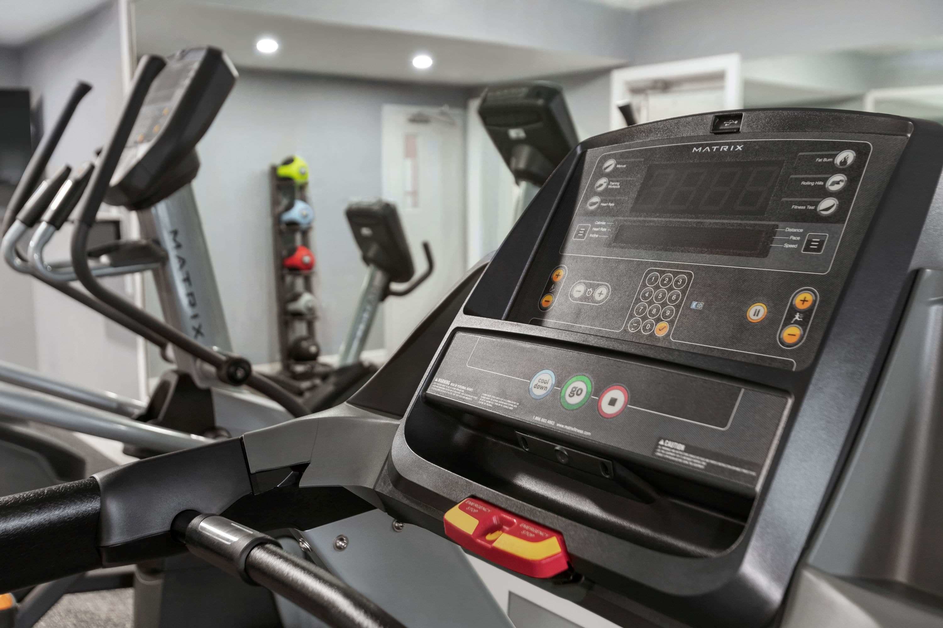 Una cinta de correr con pantalla digital y panel de control, en un gimnasio con otros equipos de ejercicio.