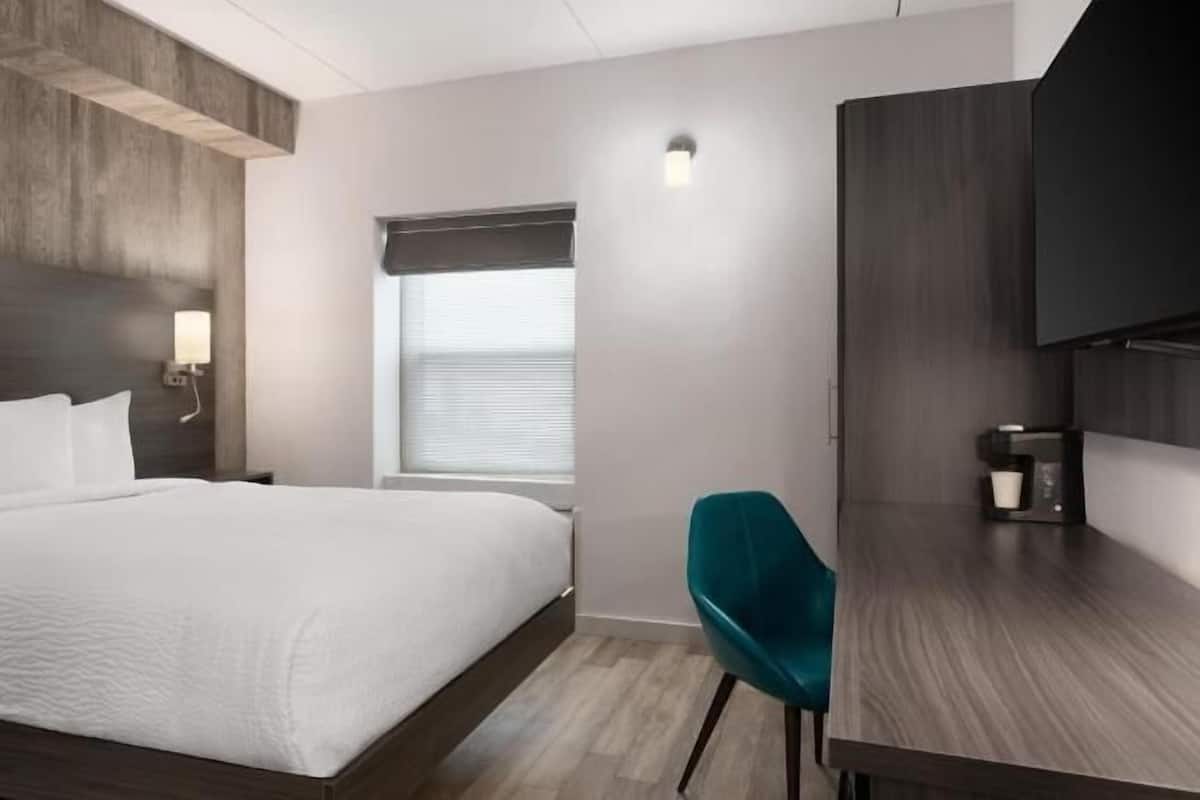 Ett modernt hotellrum med en stor säng, ett skrivbord, en stol, en TV och ett fönster med persienner.