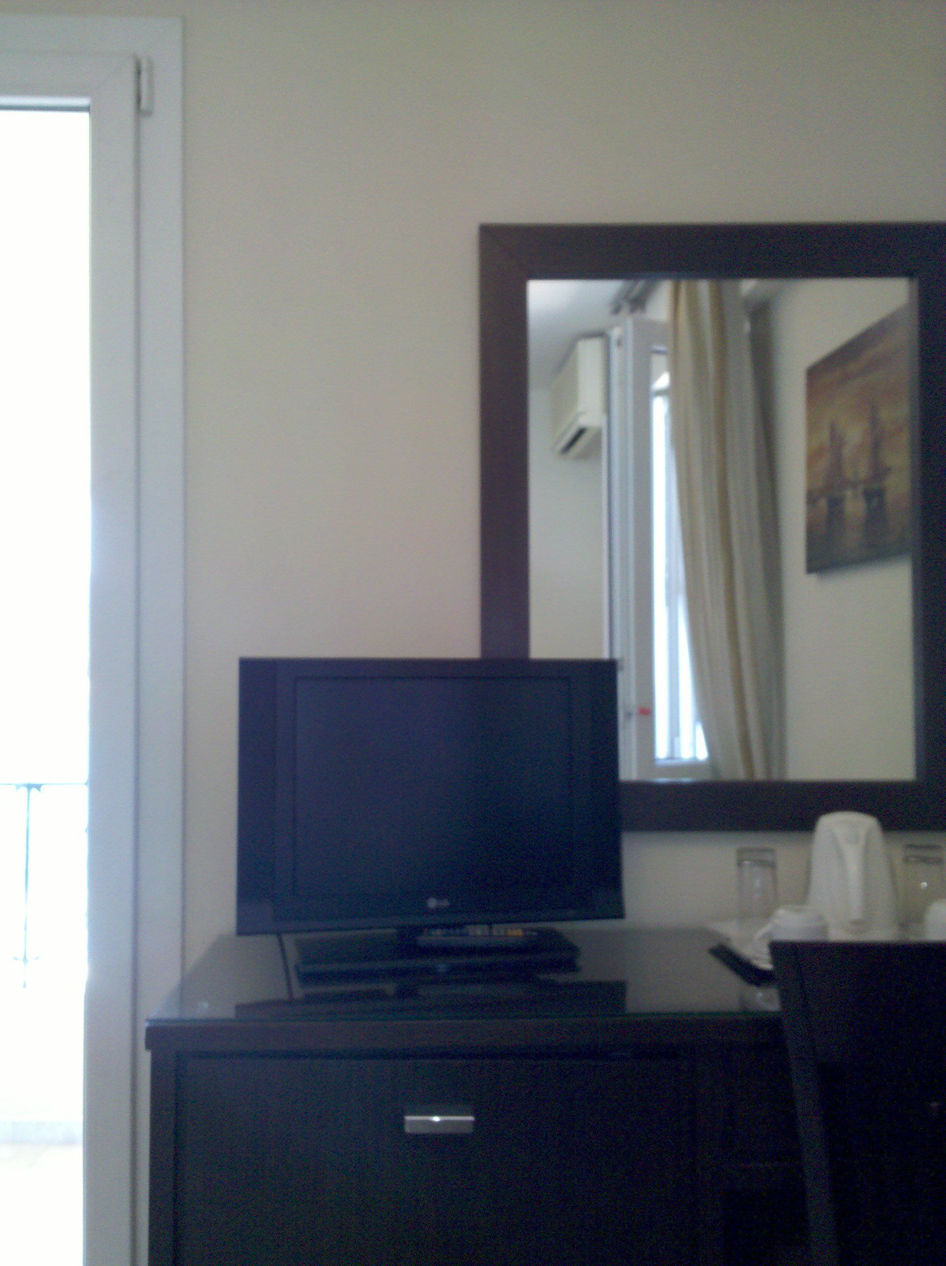 Quarto de hotel com televisão sobre uma cômoda escura, espelho na parede e janela com cortinas.