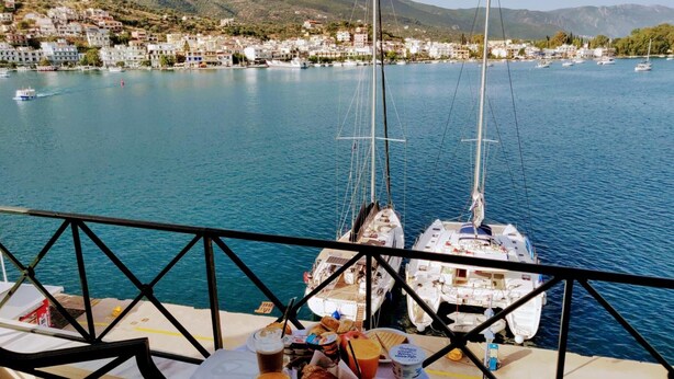 A dining table with food and drinks on a boat, overlooking a marina with sailboats and a coastal town in the background. - Manessi City Hotel (Poros)