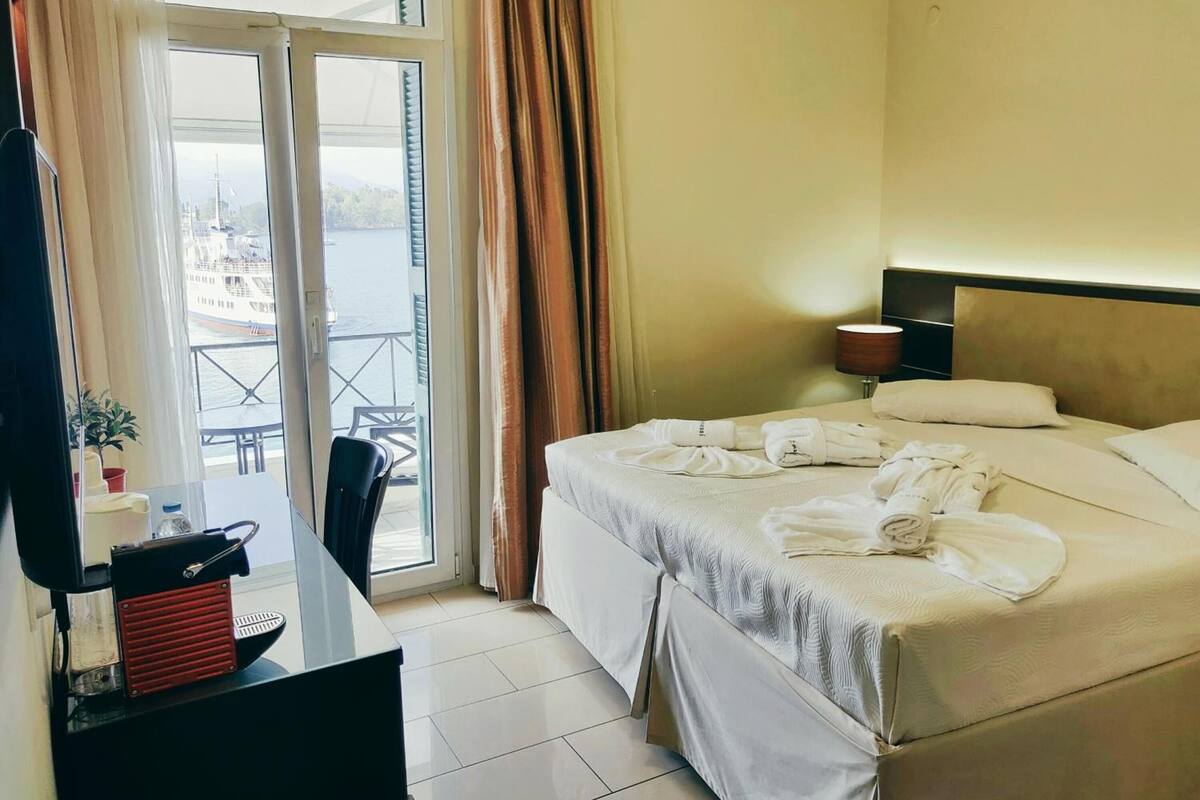 A hotel room with a bed, a desk with a red and black radio, a lamp, and a view of a body of water through the window.