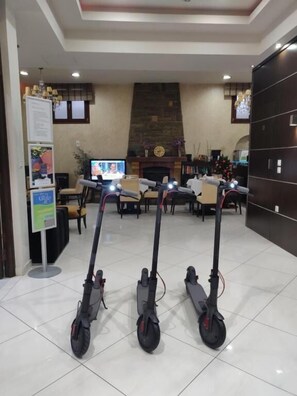 Three electric scooters parked in a lobby with a fireplace, a clock, and a television displaying a news program. - Manessi City Hotel (Poros)
