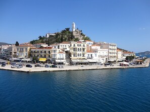 A coastal town with a prominent white building featuring balconies and a bell tower, surrounded by other buildings and a marina. - Manessi City Hotel (Poros)