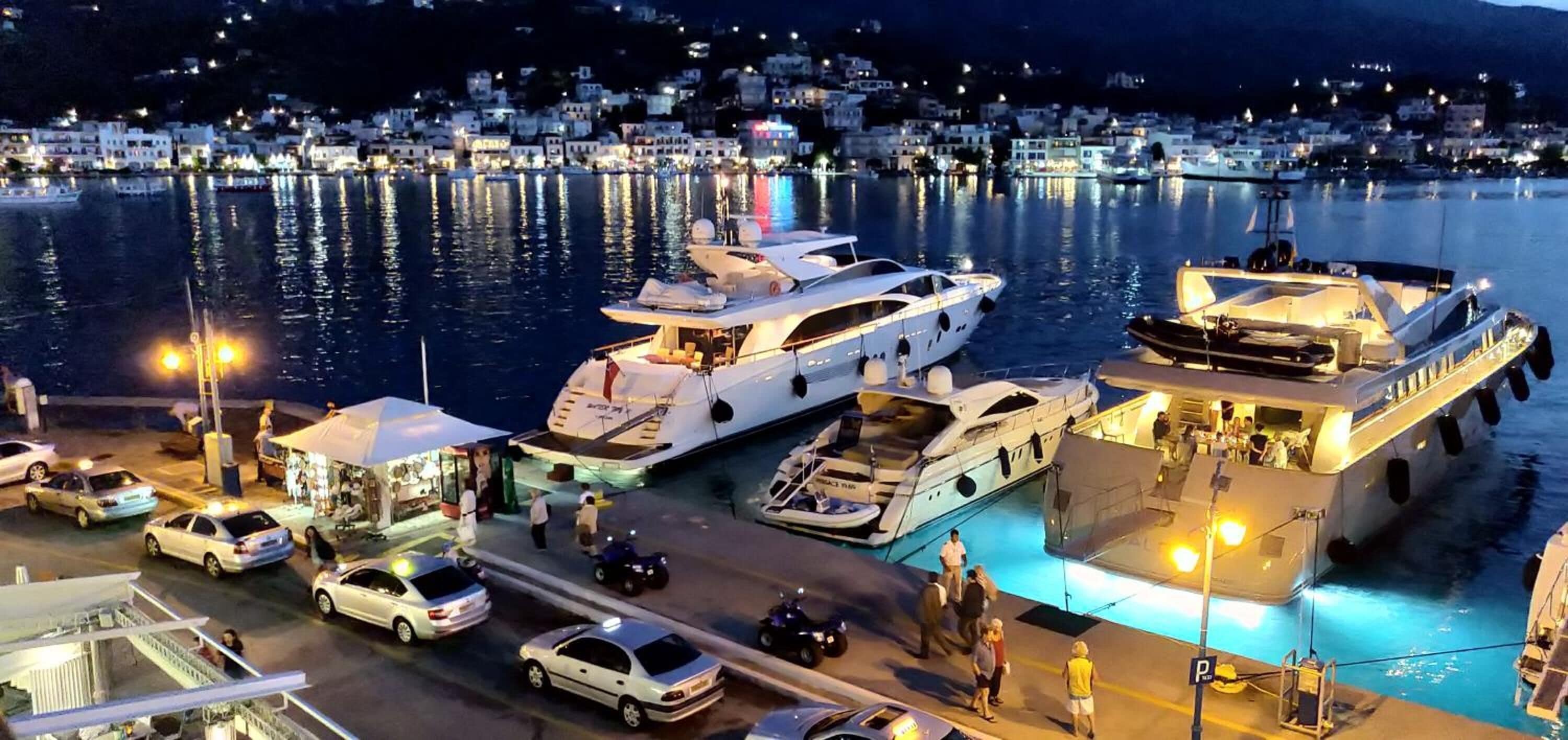 Un puerto deportivo con varios yates amarrados, un bote pequeño y algunos coches en el muelle.