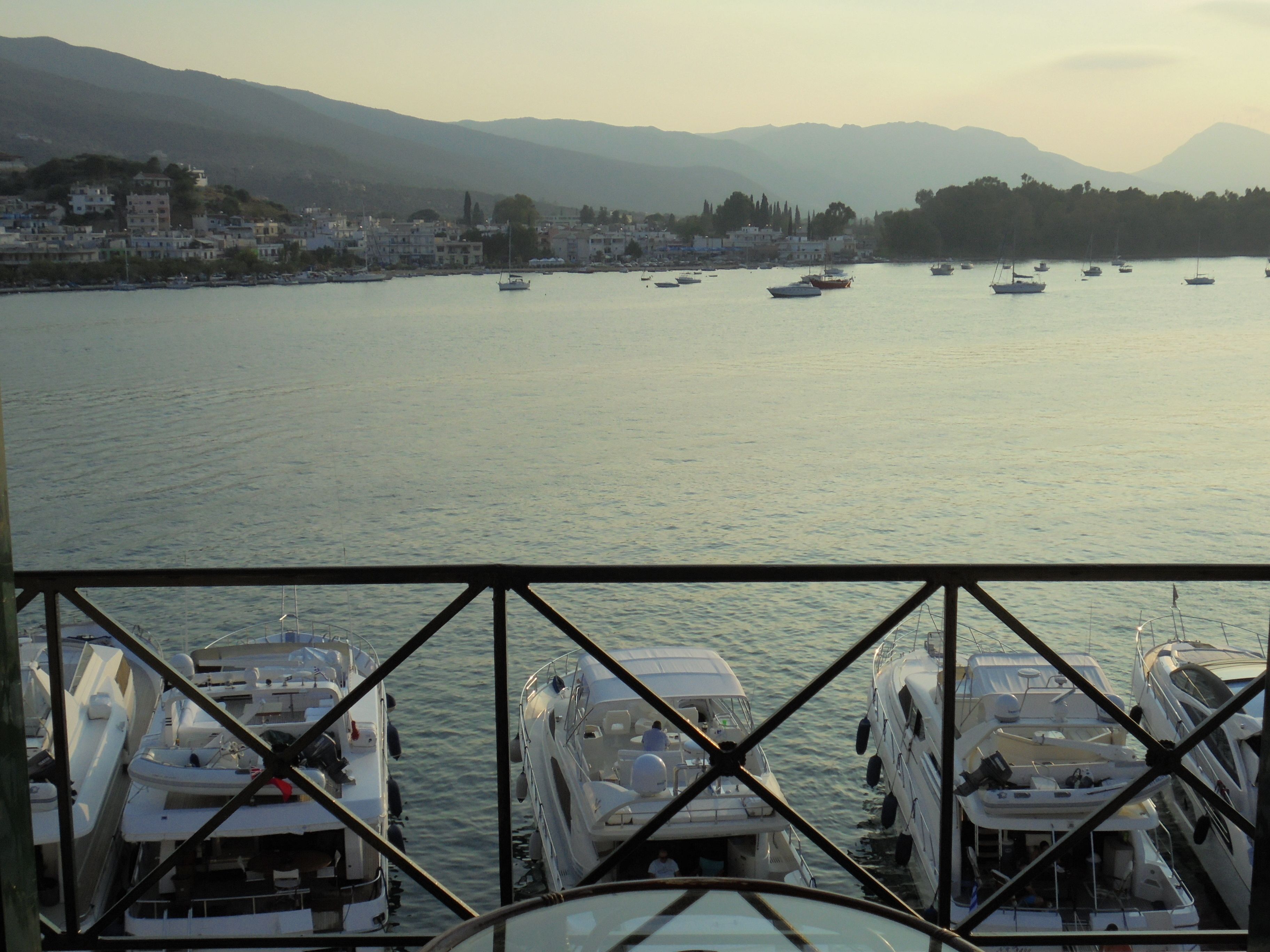 A marina with several boats docked, a view of the water, and a distant town with buildings and mountains.