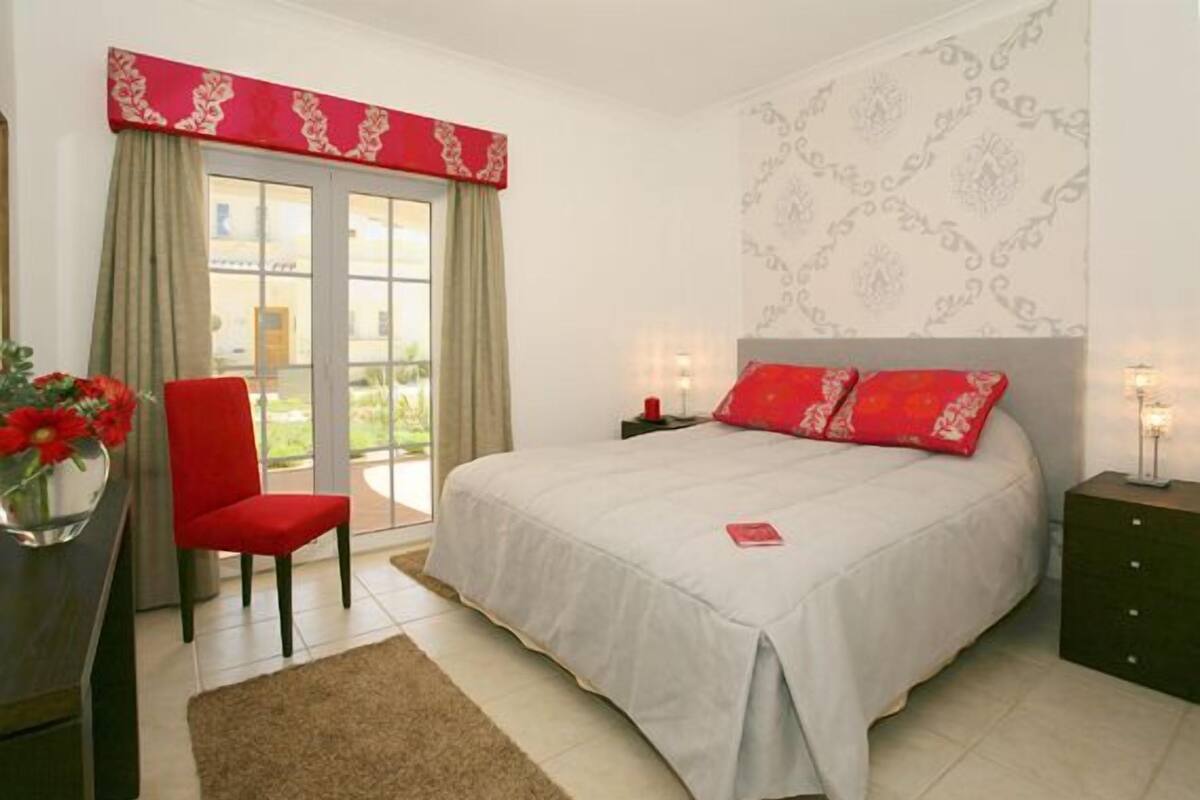 A bedroom with a bed, a chair, a nightstand, and a window with curtains.