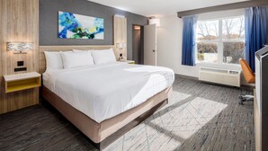 Habitación de hotel con una cama grande, cabecera de madera, un cuadro abstracto en la pared, mesita de noche con lámpara y ventana con cortinas.