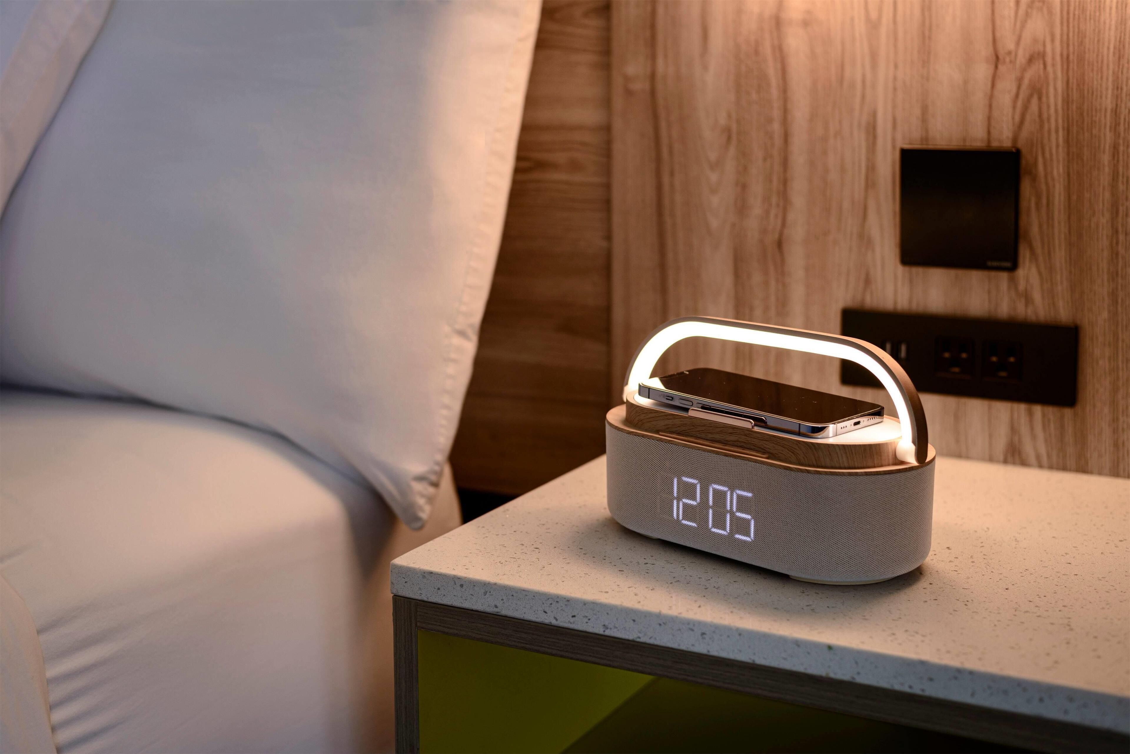 A bedside table with a digital clock displaying 12:05, a smartphone charging, and a wall-mounted control panel.