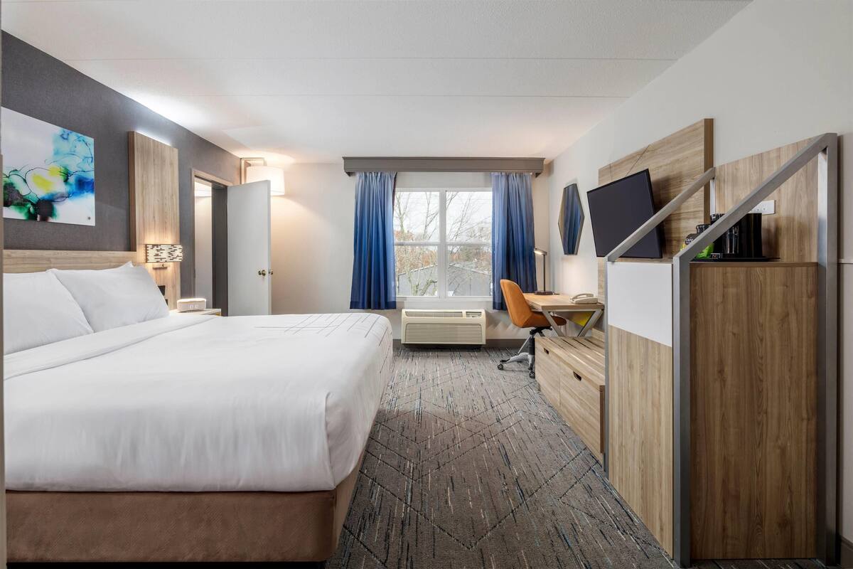 A hotel room with a large bed, a desk with a chair, a flat-screen TV, and a window with curtains.