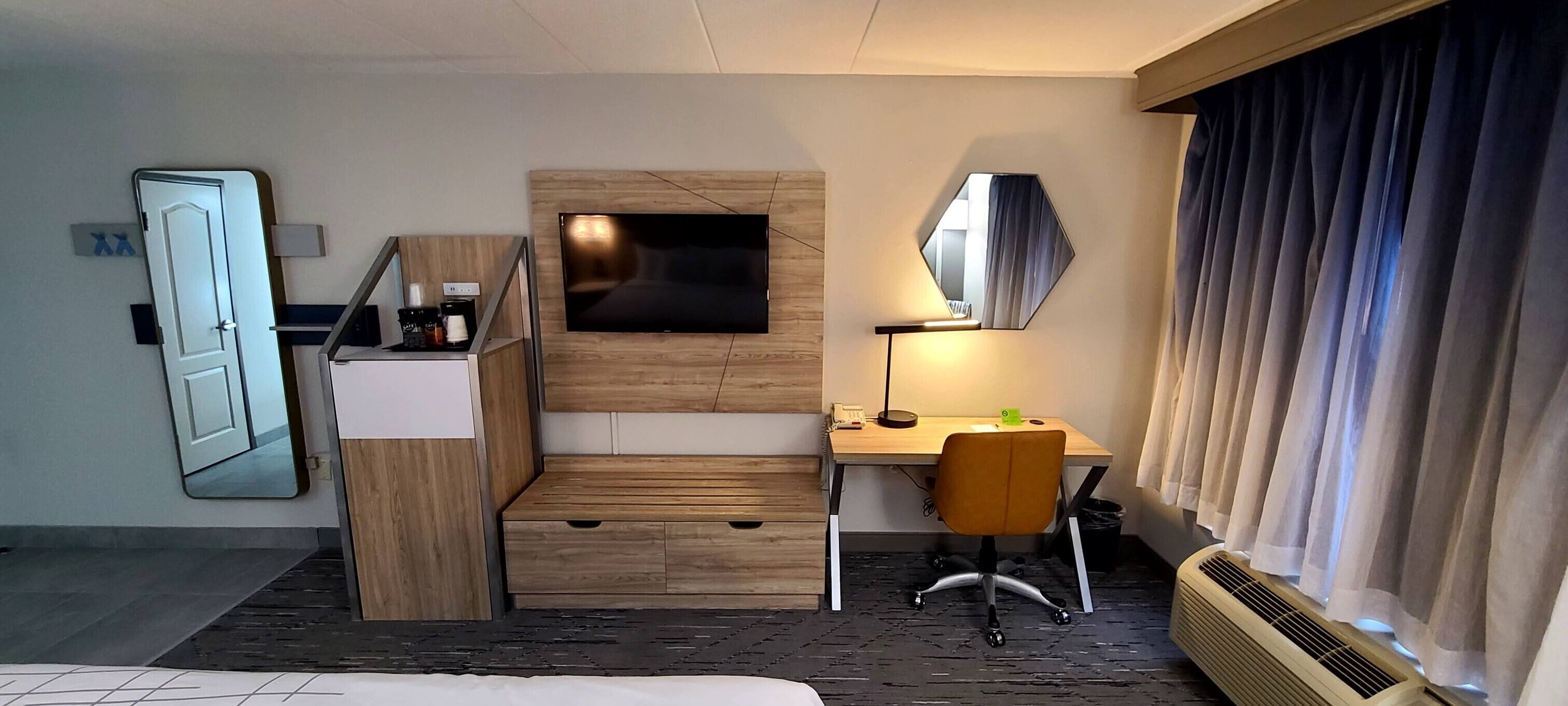A hotel room with a bed, a desk with a chair, a TV, a mirror, a heater, and a window with curtains.