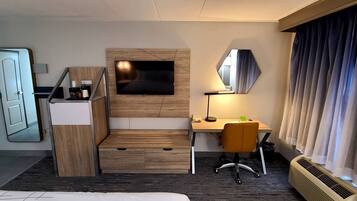 A hotel room with a bed, a desk with a chair, a TV, a mirror, a heater, and a window with curtains.
