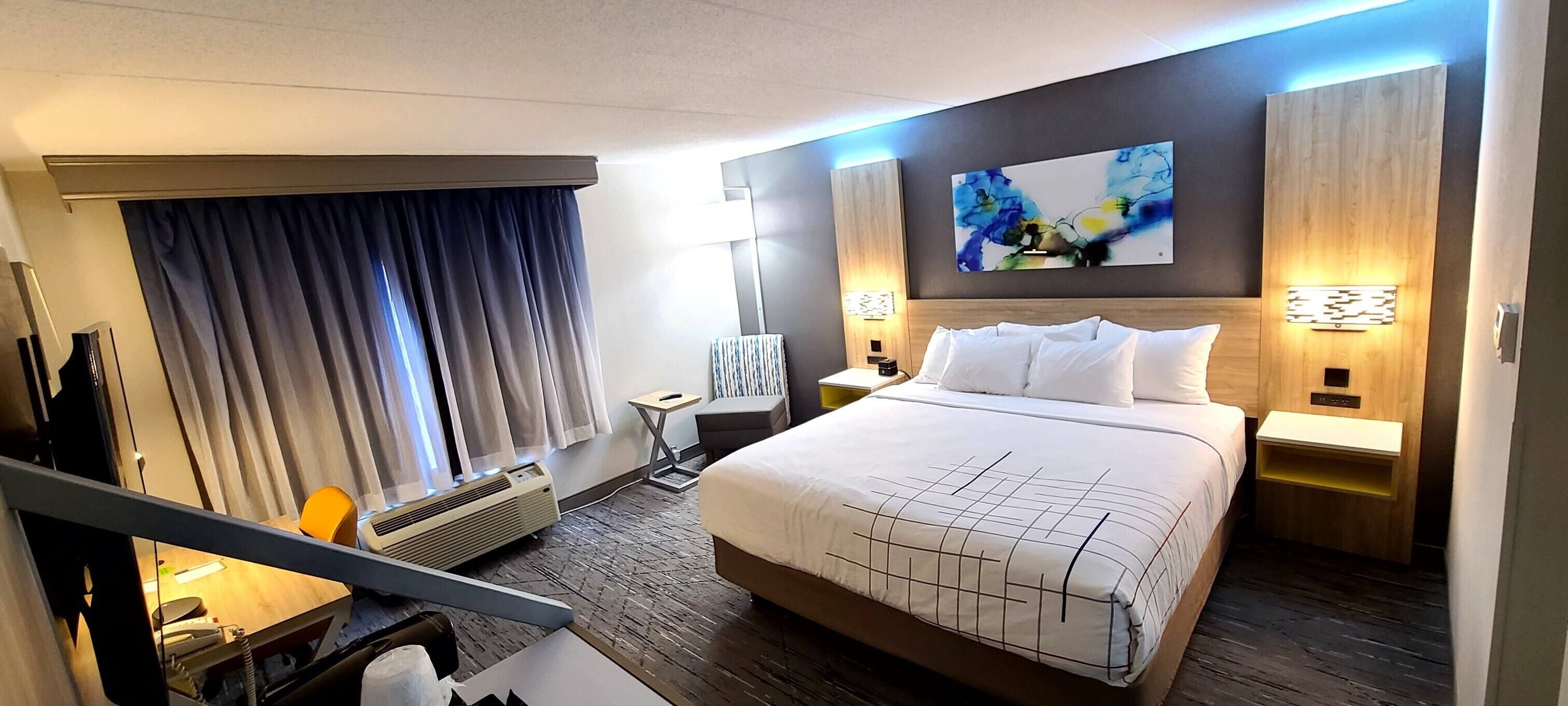 A hotel room with a large bed, a desk with a chair, a desk lamp, a window with curtains, and a wall-mounted artwork.
