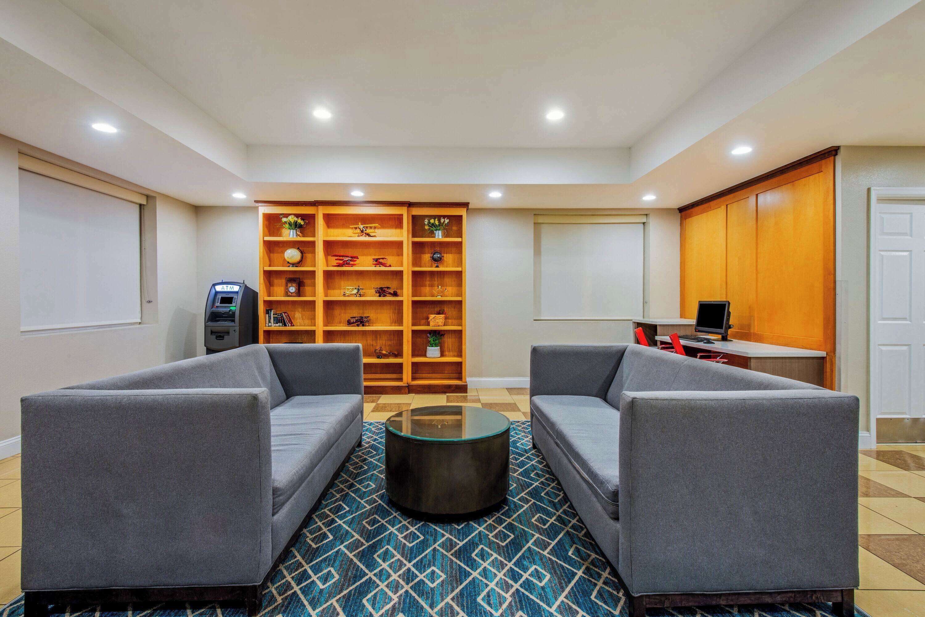 A lobby with two grey sofas, a coffee table, a wooden bookshelf, a desk with a computer, and a vending machine.