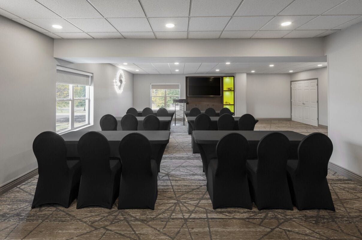 Un salón de conferencias con varias mesas y sillas, una pantalla grande y una puerta con detalles en verde.