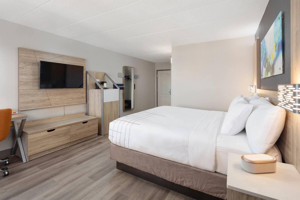 A modern hotel room with a large bed, a desk with a chair, a TV mounted on the wall, and a small bedside table with a lamp.