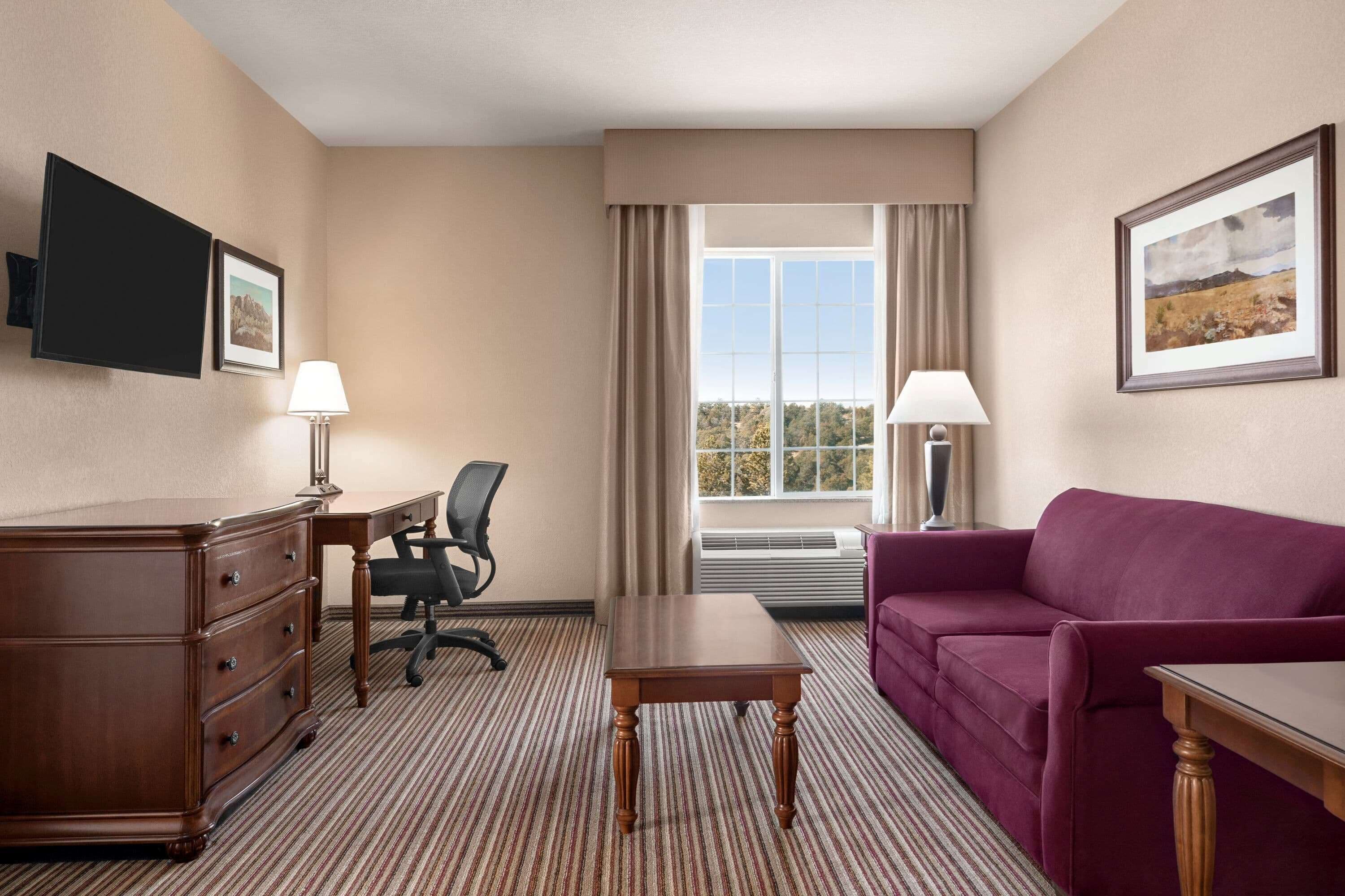 A hotel room with a wooden dresser, a desk, a purple sofa, a small table, a chair, and a large window with curtains.