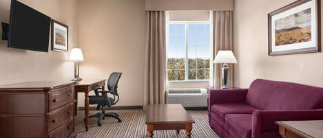 A hotel room with a wooden dresser, a desk, a purple sofa, a small table, a chair, and a large window with curtains.