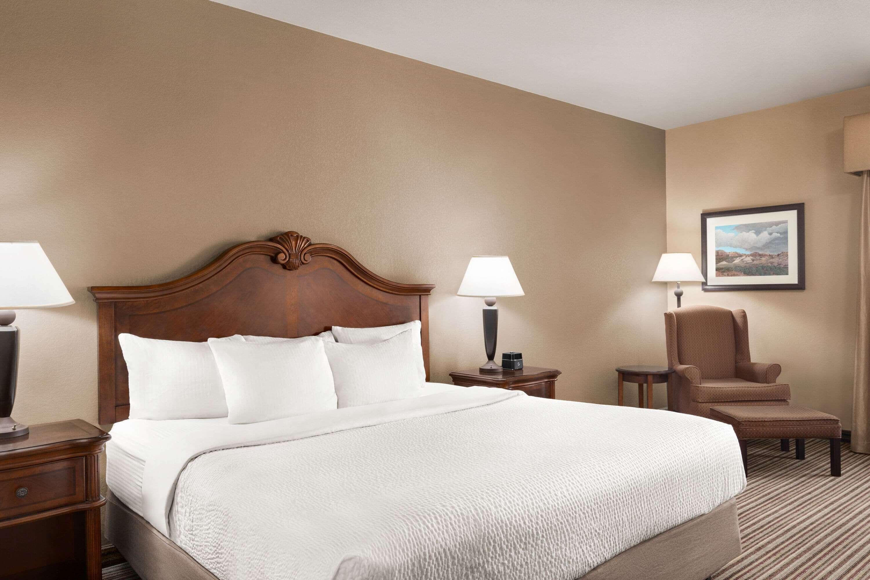 Una habitación de hotel con una cama grande, dos mesitas de noche, una silla y una lámpara.
