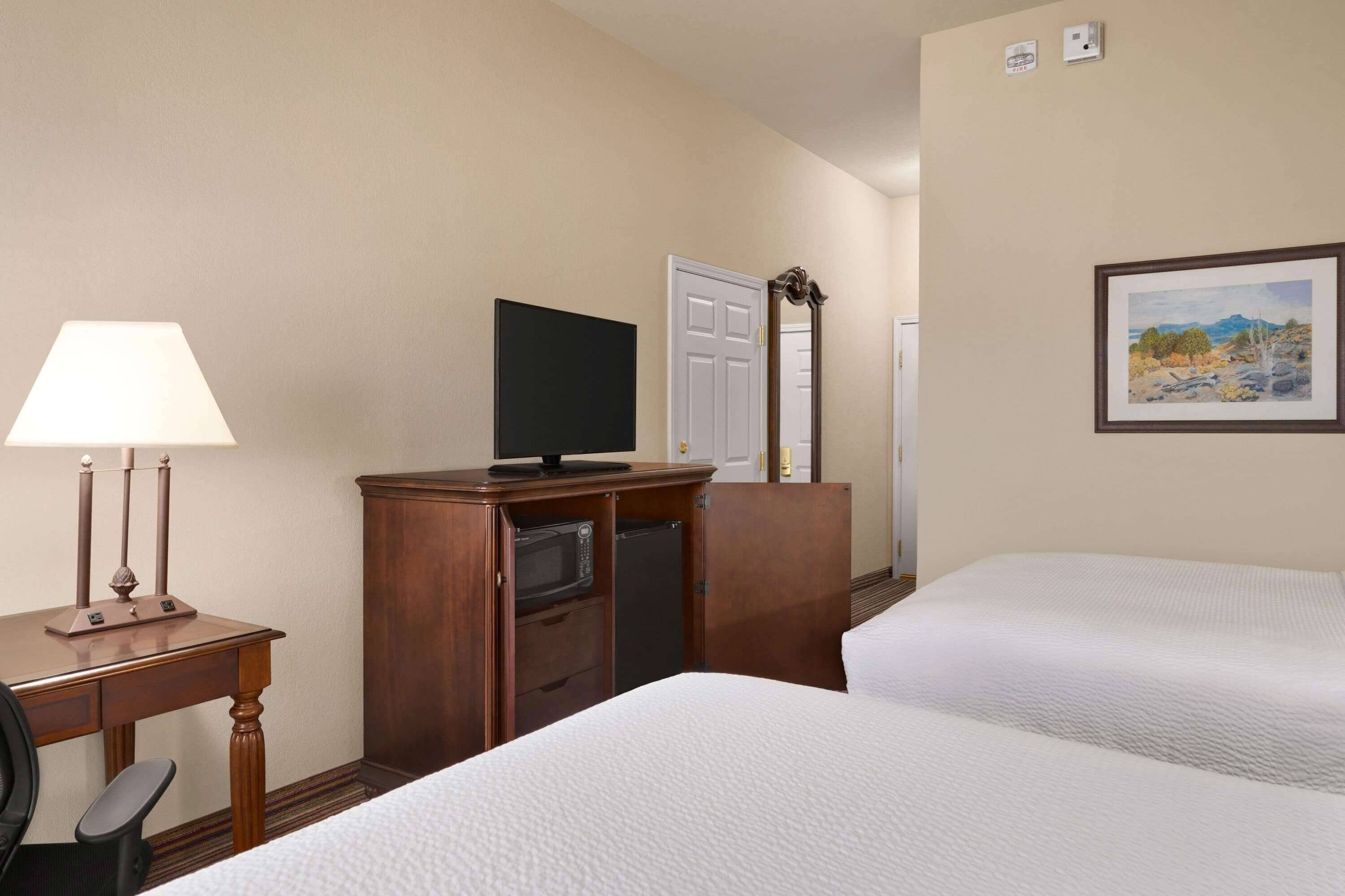 A hotel room with two beds, a desk with a lamp, a television, a microwave, and a painting on the wall.