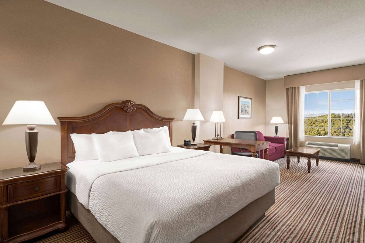 Een hotelkamer met een groot bed, twee nachtkastjes met lampen, een bureau met stoel, en uitzicht op een bergachtig landschap.