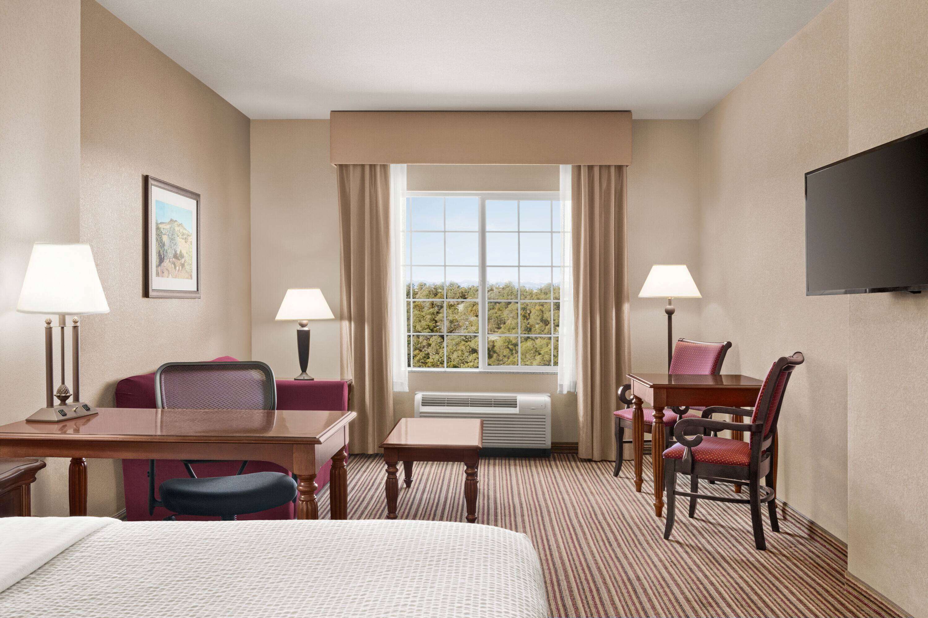 A hotel room with a bed, two chairs, a table, a TV, and a window with curtains.