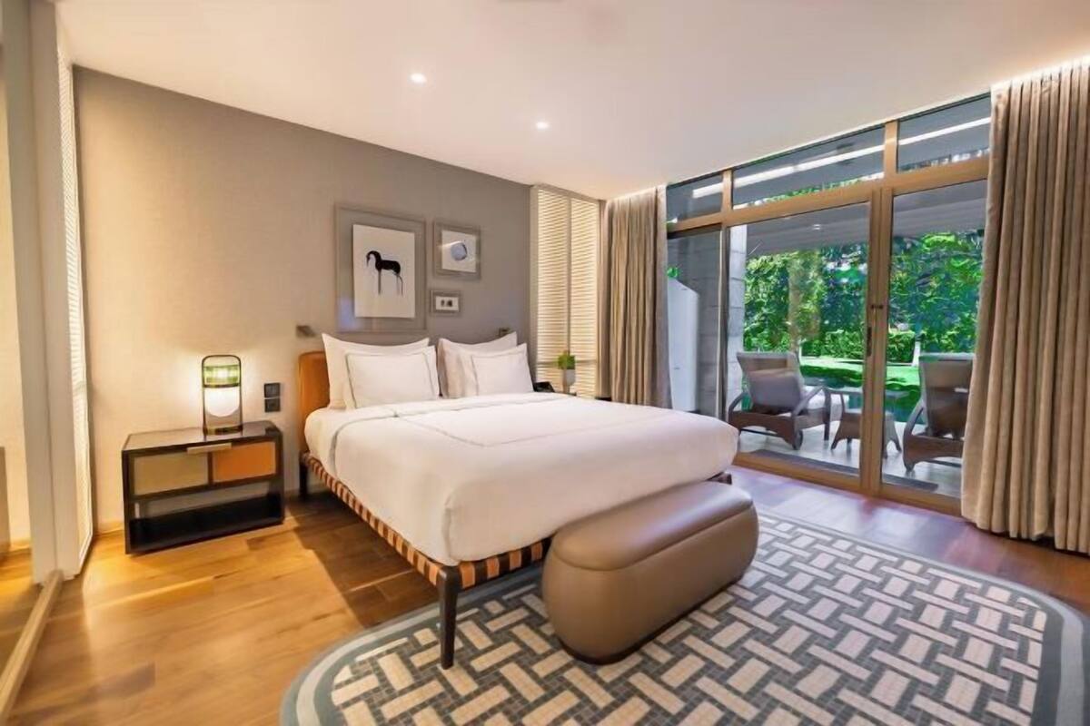 A modern hotel room with a large bed, a bedside table, a lamp, and a view of the outdoors through a glass door.