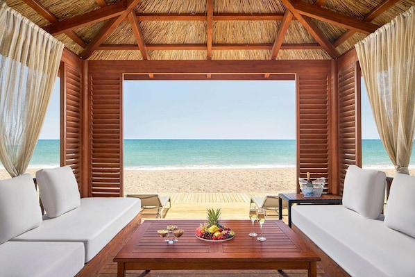 A beachside room with a wooden ceiling, two white sofas, a wooden table with fruit and drinks, and a view of the ocean.