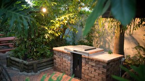 Un espace barbecue en briques avec un gril intégré, entouré d’une végétation luxuriante et agrémenté d’un banc en bois.