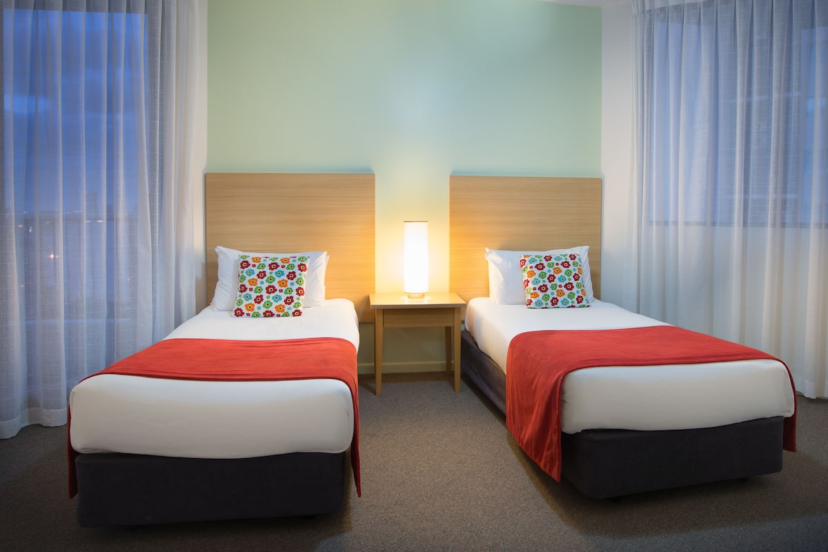 Quarto de hotel com duas camas, cada uma com um cobertor vermelho, roupa de cama branca e almofadas florais coloridas. Há uma mesinha de cabeceira com uma luminária entre as camas.